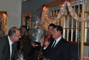 man speaking into microphone while man next to him holds up silver bucket