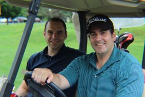 two men sitting in golf cart smiles for camera