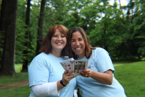 two women in light blue aim services shirts smiling while holding playing cards