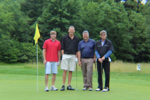 group of four golfers smiling on green next to yellow pin