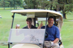 men in golf cart smiling, giving thumbs up and toasting beer