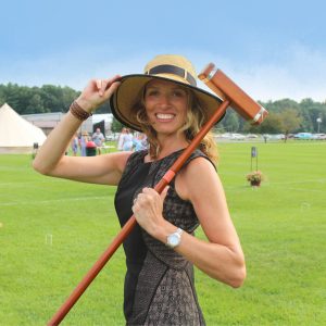 Woman standing in grassy field holding brim of sun hat and croquet mallet over her shoulder smiling