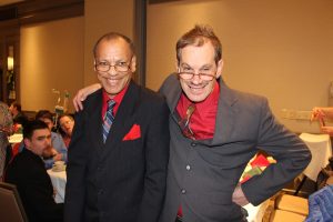 two men in suits and glasses smile with one another while posing for picture