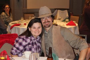 man with cowboy hat and vest smiling with arm around woman in purple flannel