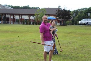 man in purple polo holds croquet mallet and baby while smiling