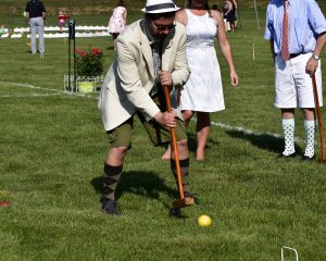 man in hat swings mallet like a pendulum at croquet ball