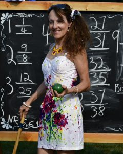 woman in floral dress stands in front of results chalk board displaying green croquet ball