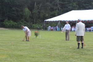 two men in white watching man smack red croquet ball