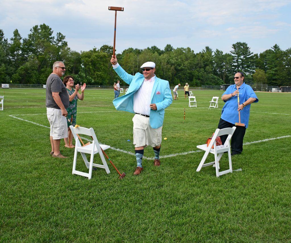 Man in teal jacket and white hat holding croquet mallet in the air at Croquet on the Green 2019