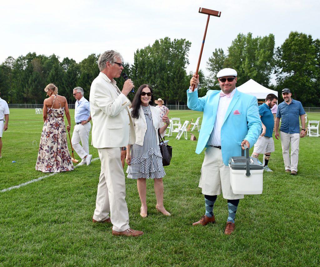Man in teal coat and white hat holding mallet in the air with a cooler in the other hand as Walt Adams speaks into a microphone at Croquet on the Green 2019