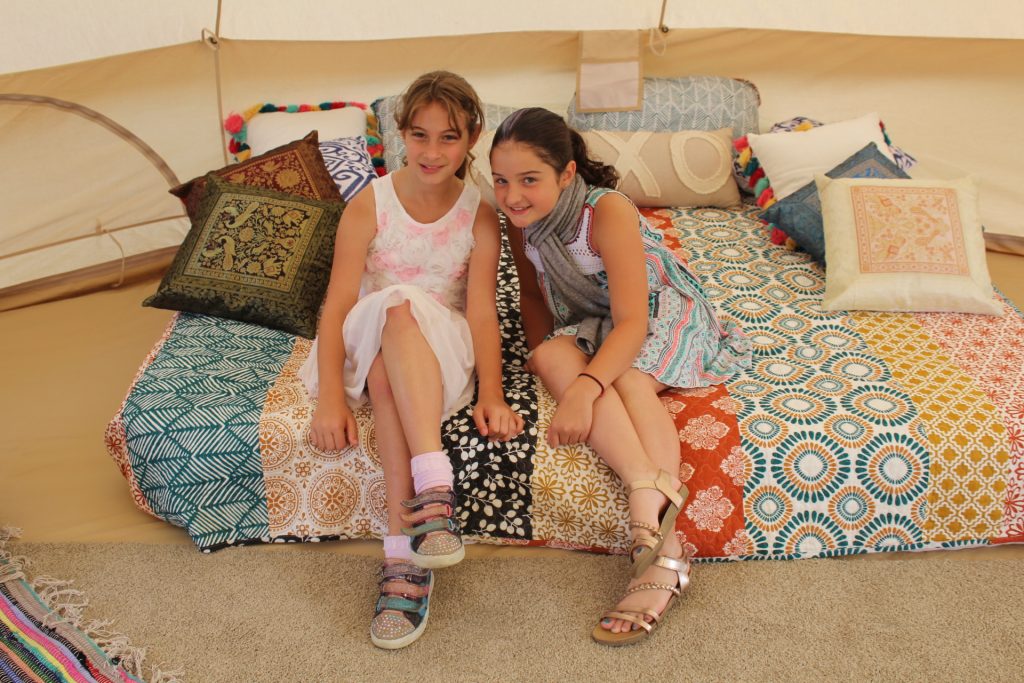 Two young girls sitting on a bed with a patchwork pattern blanket in the glamping tent at Croquet on the Green 2019