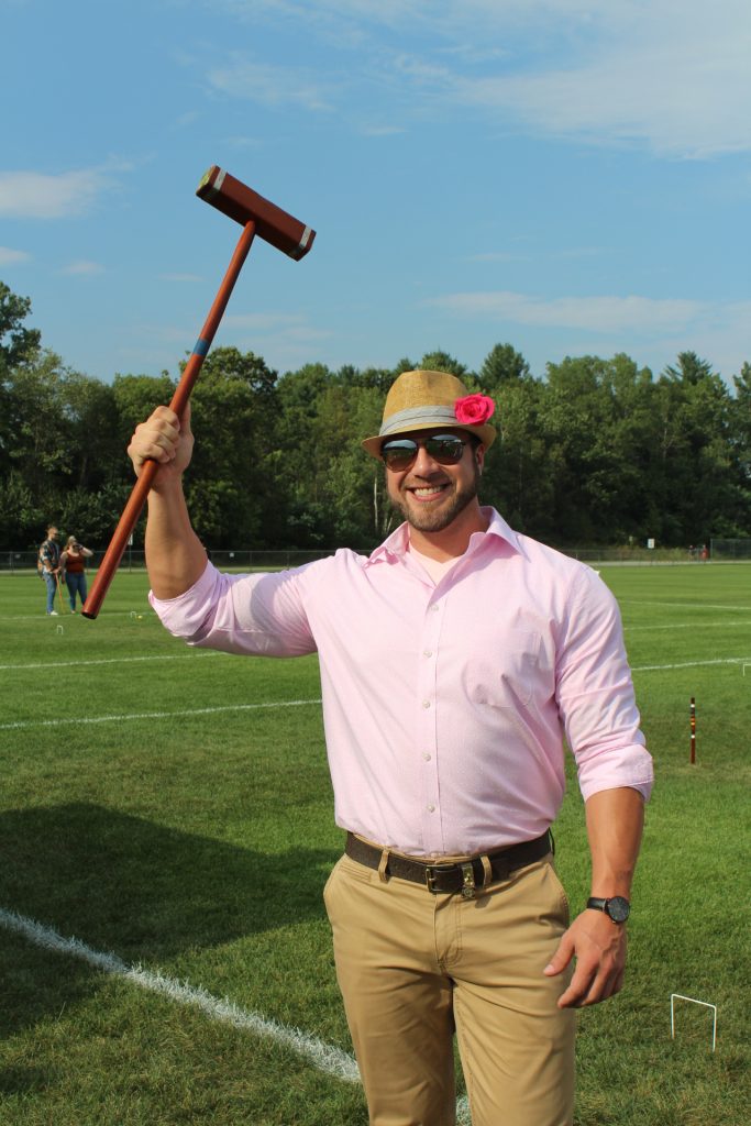 Man in pink shirt with tan fedora and a pink flower in the hat holding a croquet mallet in the air and smiling at Croquet on the Green 2019
