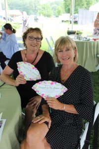 Two women each holding a fan that says "Croquet on the Green" sitting at a table