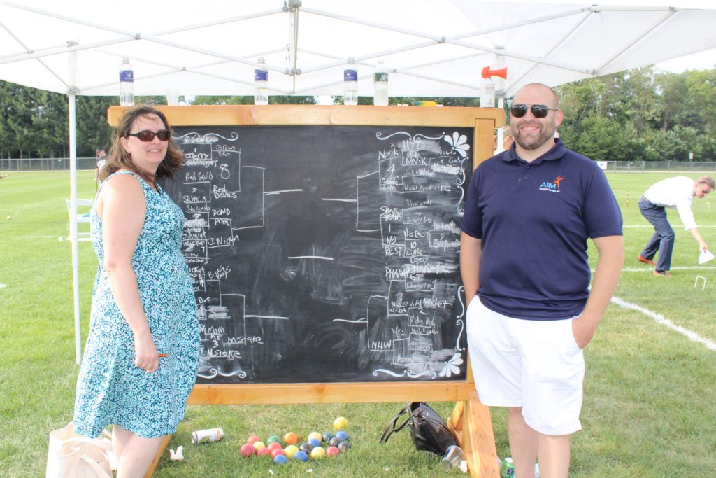 Two people standing in front of a large chalkboard sign that has a bracket of team names on it at Croquet on the Green 2019
