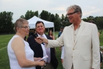 June MacClelland speaking into a microphone held by Walt Adams as Chris Lyons looks on smiling at Croquet on the Green 2019