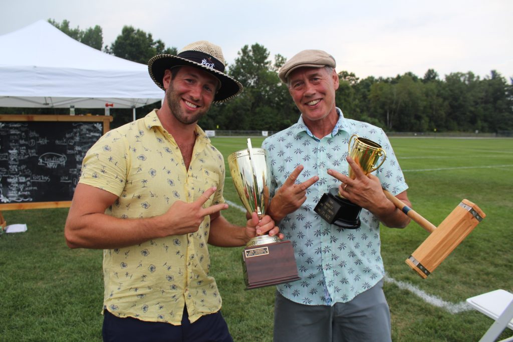 Croquet winners, Brett and Brett Jr. Armstrong, holding two trophies, a croquet mallet, and two fingers up at Croquet on the Green 2019