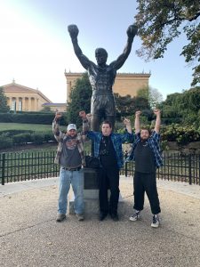Three man standing in front of the Rocky statue in Philadelphia, PA with their hands all up in the air
