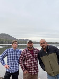 Three men standing together in front of a lake