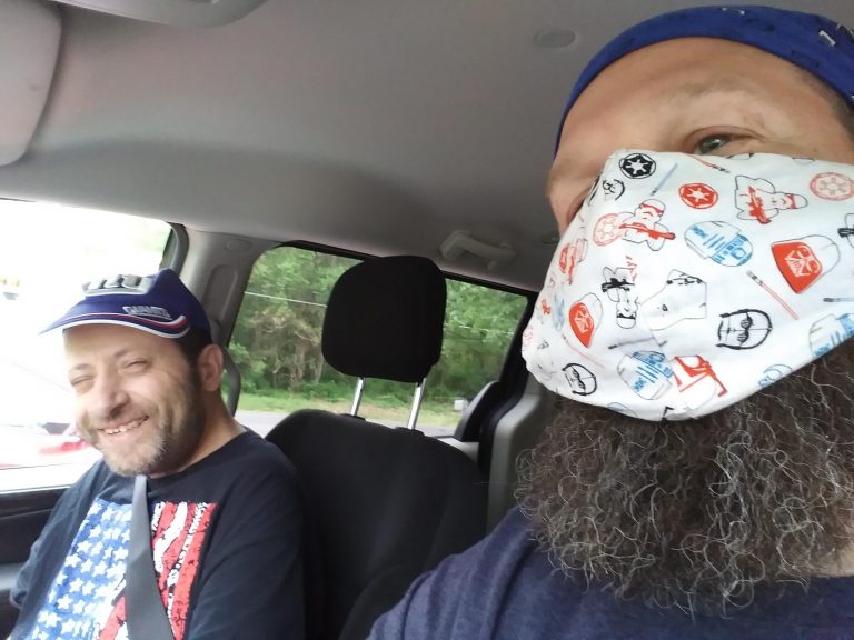 Two men sitting in a car, one with a big beard wearing a mask and another with a hat smiling