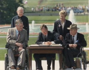 President Bush sitting at a table signing papers with four people sitting around him, two in wheelchairs
