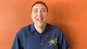 Andrew in a blue button up shirt with a palm tree on it standing in front of a bright orang wall.