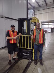 Two people in orange vests standing in front of a fork lift
