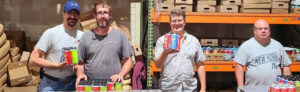 Four people standing together holding canned drinks in front of shelves with many cardboard boxes on them